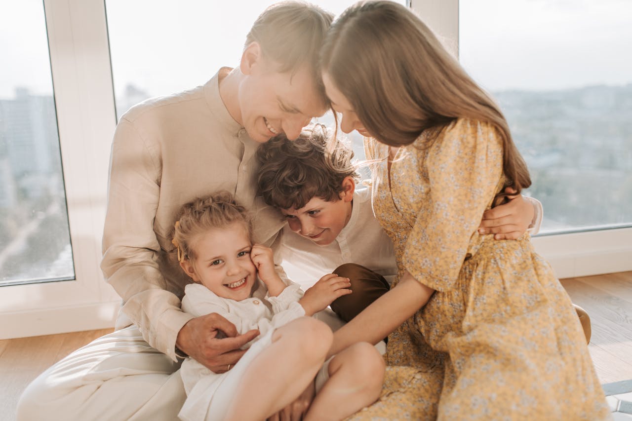 Cheerful family of four cuddling and smiling together in a bright, sunlit living room setting.
