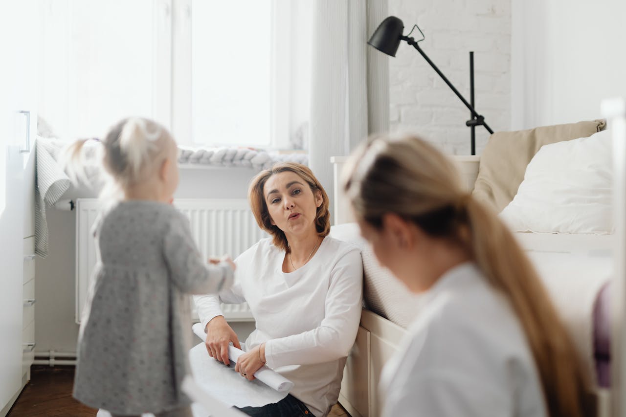A mother sits on the floor talking with her daughter in a cozy bedroom setting.